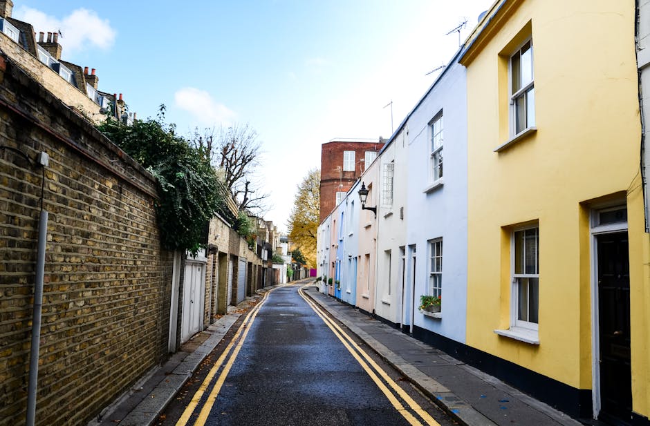 A narrow residential street with a gently curved asphalt road lined by brick houses with pitched roofs, some featuring chimneys. Tall, leafless trees on both sides cast over the scene, with branches e